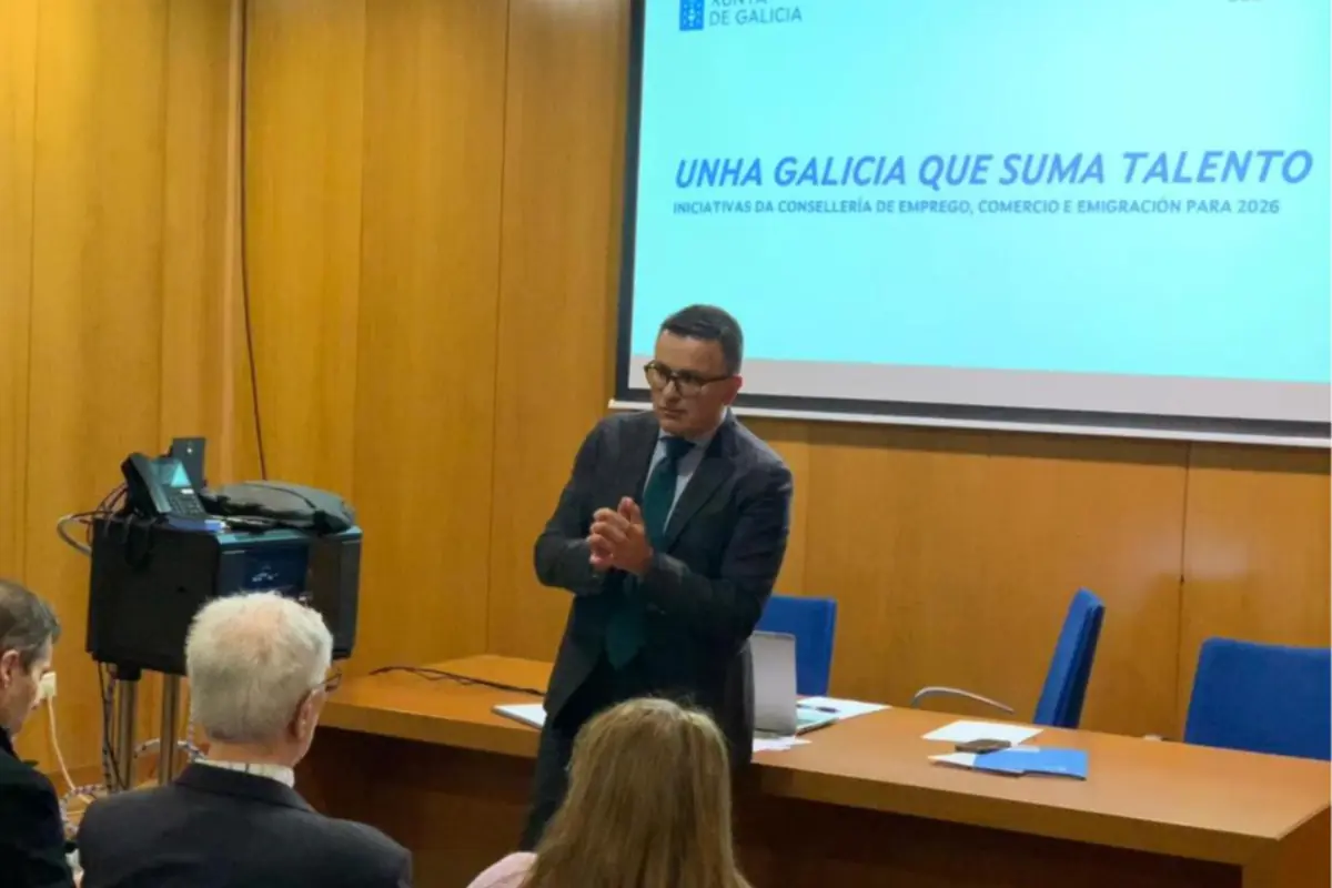 Hombre con traje dando una charla en una sala de conferencias frente a un proyector con la frase Unha Galicia que suma talento mientras varias personas lo escuchan sentadas