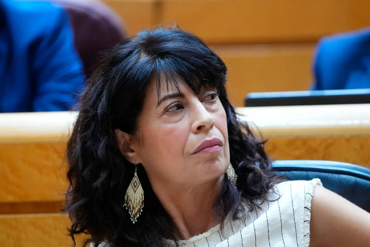 Mujer de mediana edad con el pelo oscuro y rizado sentada en un escaño mirando hacia un lado con expresión seria y llevando pendientes largos de abalorios Mujer de mediana edad con el pelo oscuro y rizado sentada en un escaño mirando hacia un lado con expresión seria y llevando pendientes largos de abalorios