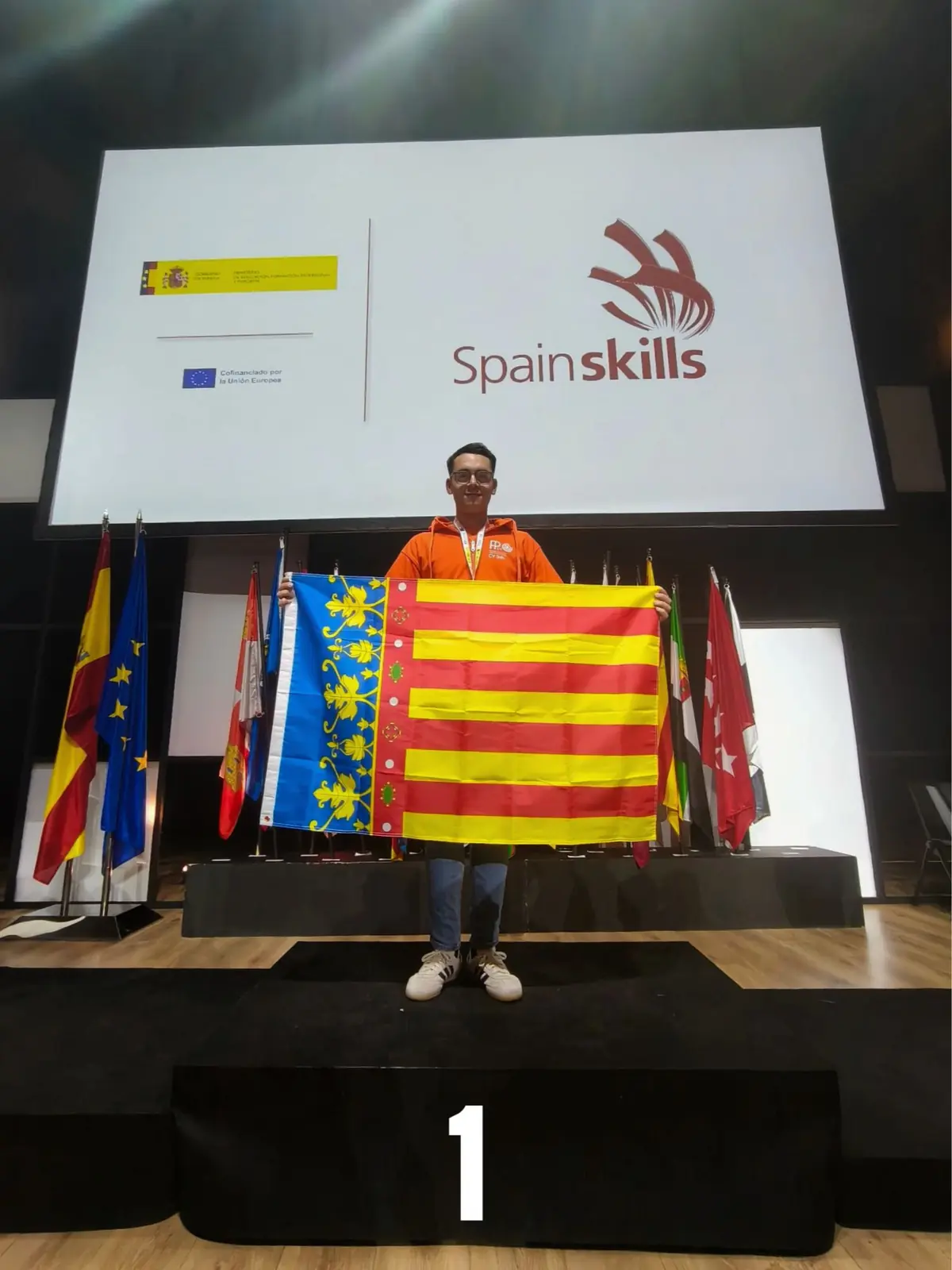 Persona joven sobre el podio en primer lugar sosteniendo una gran bandera de la Comunidad Valenciana en la ceremonia de premios de SpainSkills