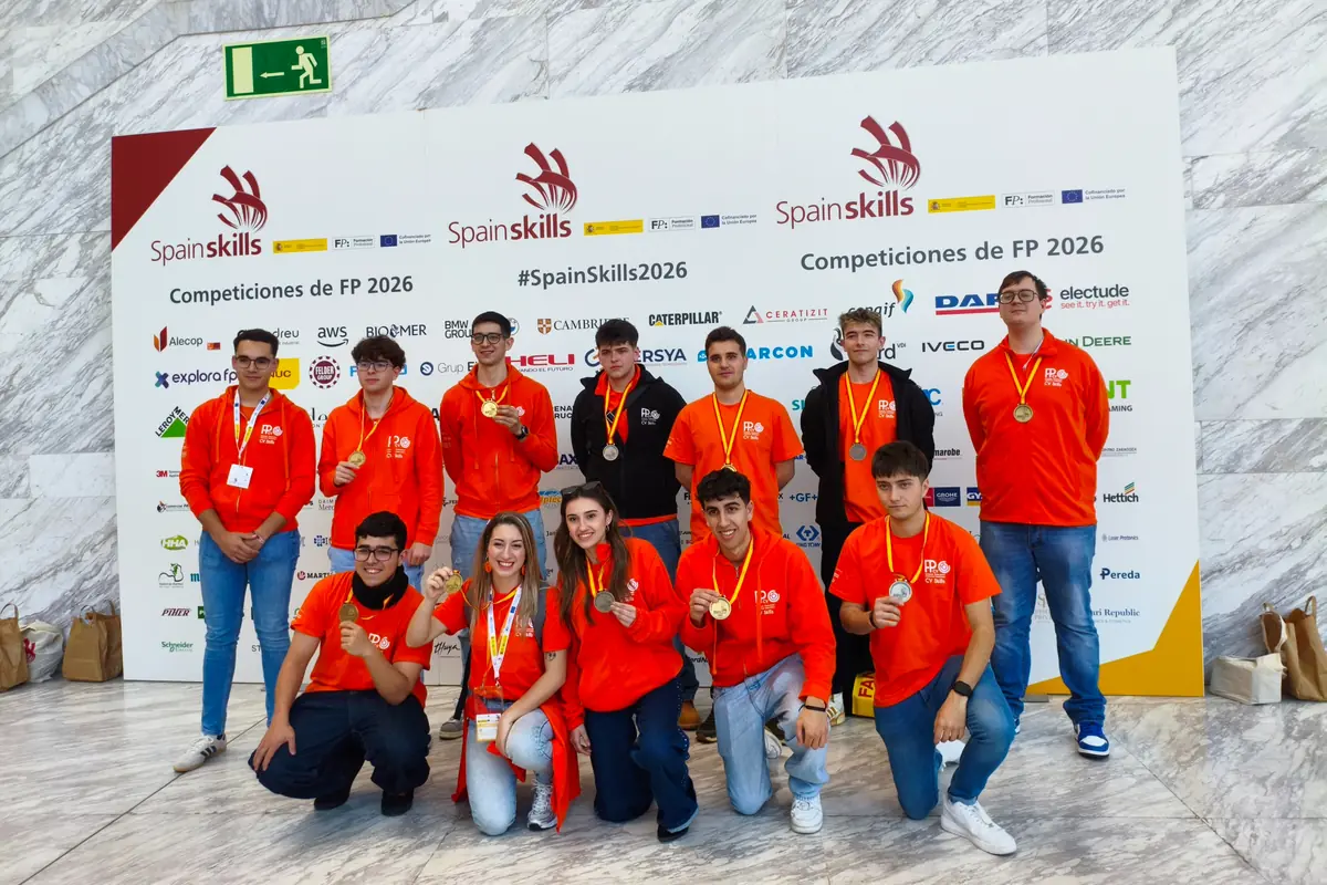 Grupo de estudiantes con sudaderas naranjas y medallas posando sonrientes frente a un panel de SpainSkills 2026 en una competición de Formación Profesional