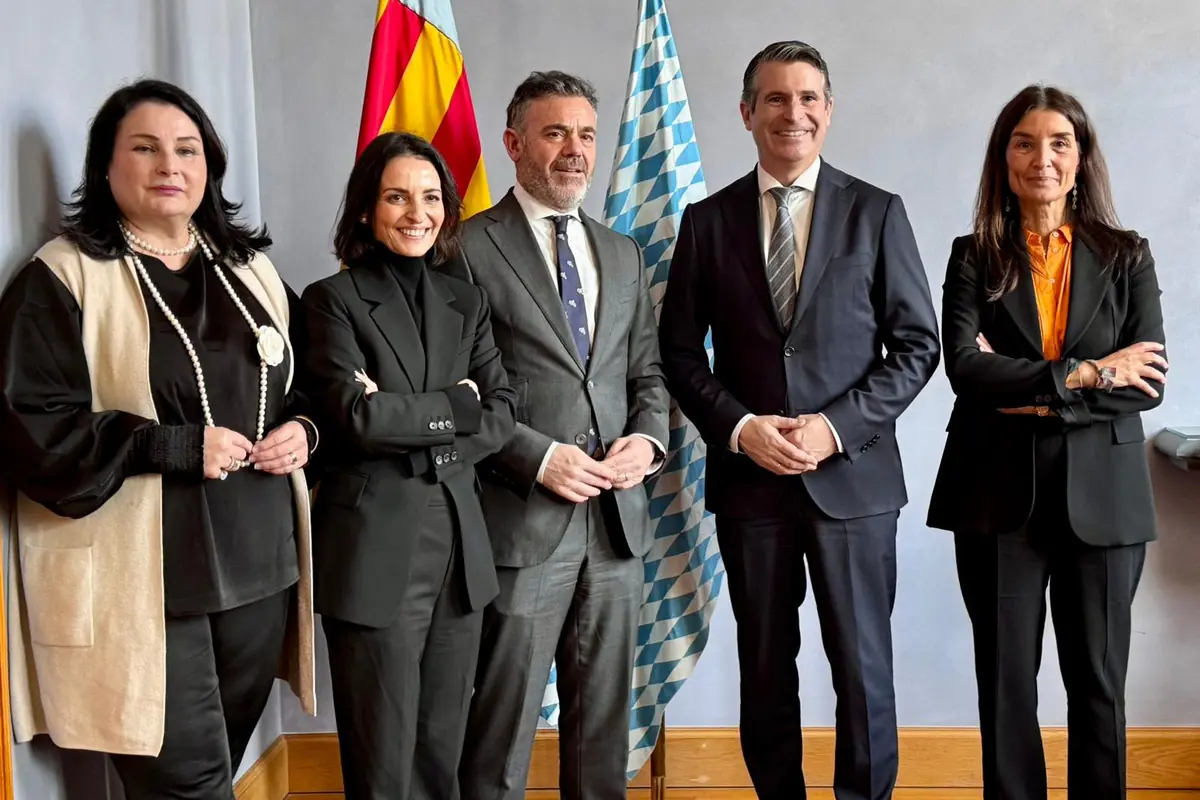 Cinco personas vestidas de forma formal posan sonrientes frente a dos banderas en un entorno institucional