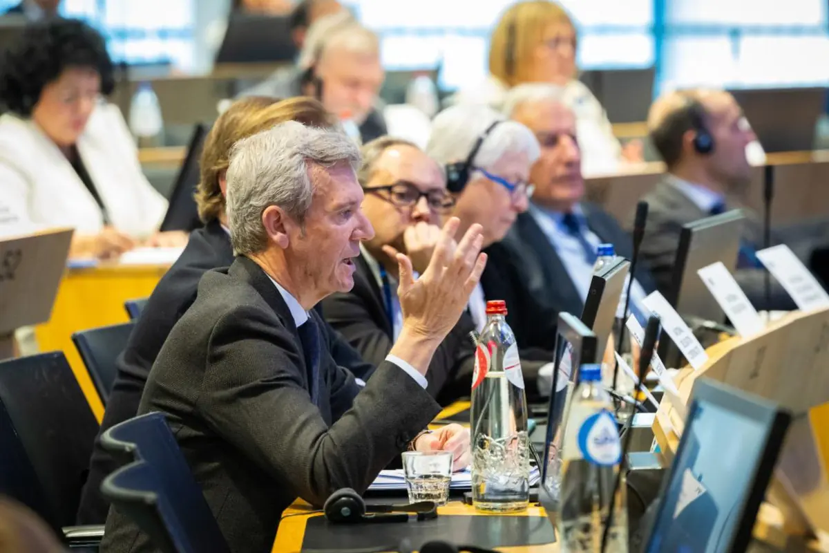 Hombre de cabello canoso interviene gesticulando durante una reunión formal en una sala de conferencias llena de personas con auriculares y ordenadores portátiles