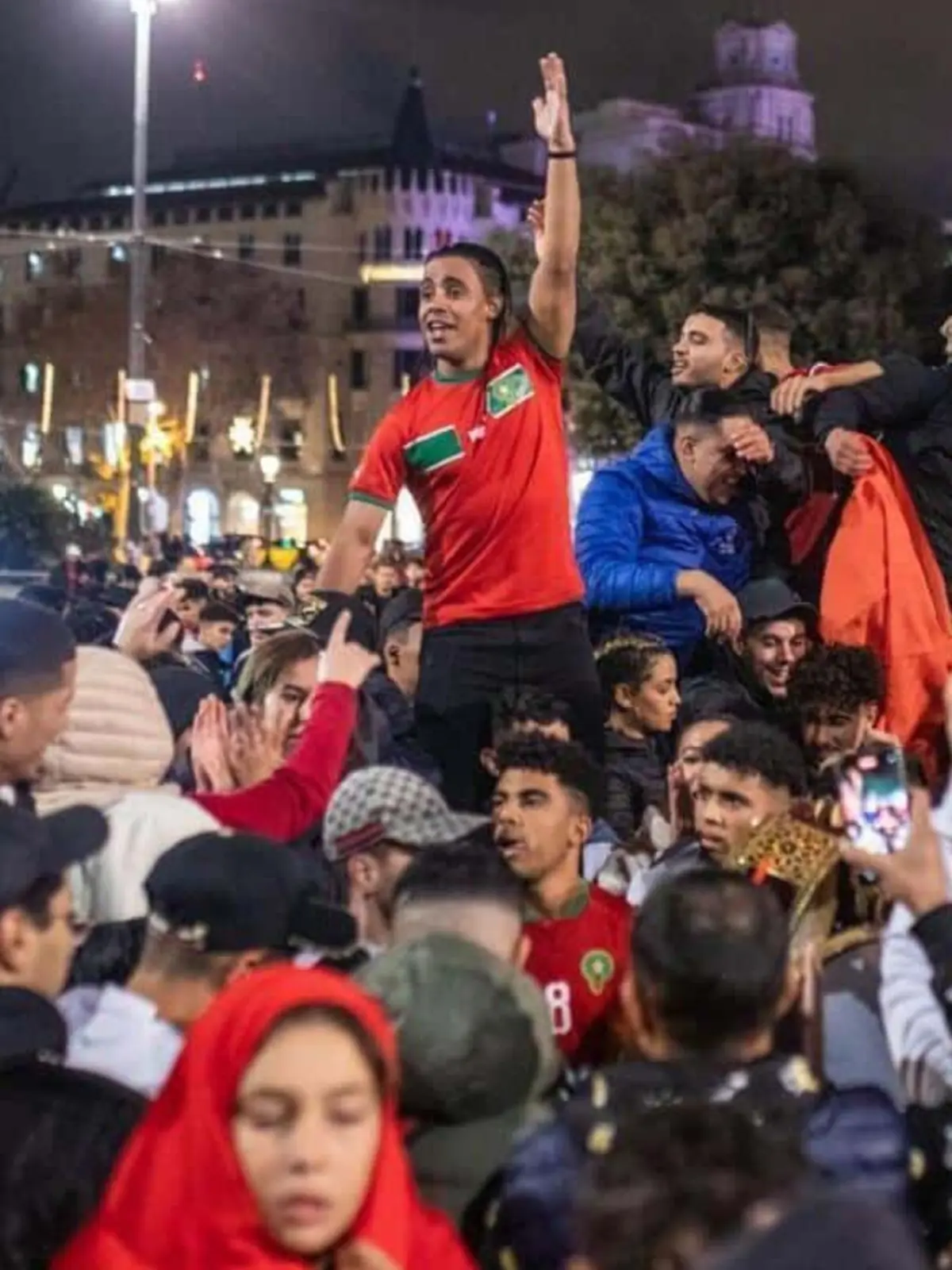 Aficionados celebrando en la calle de noche con camisetas de la selección de Marruecos mientras un joven levantado sobre la multitud saluda con el brazo en alto
