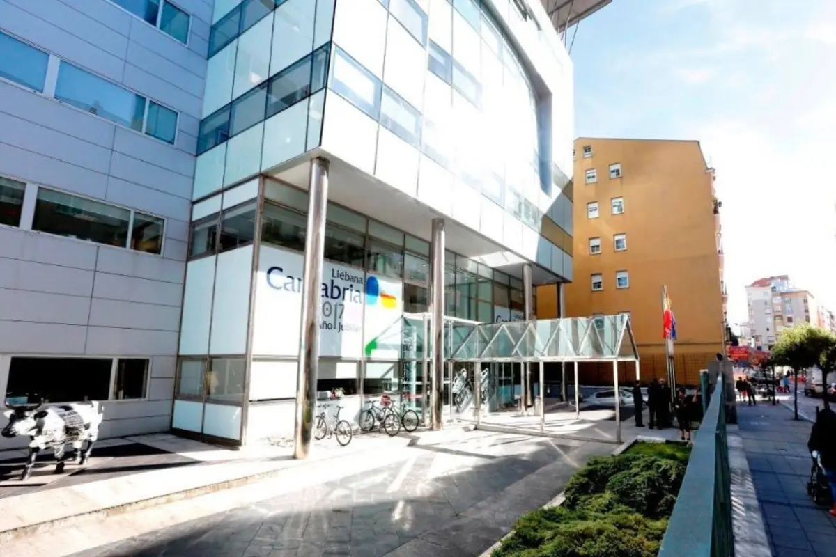 Fachada acristalada de un edificio moderno de oficinas en una calle urbana con entrada cubierta bicicletas aparcadas y varias personas junto a unas banderas