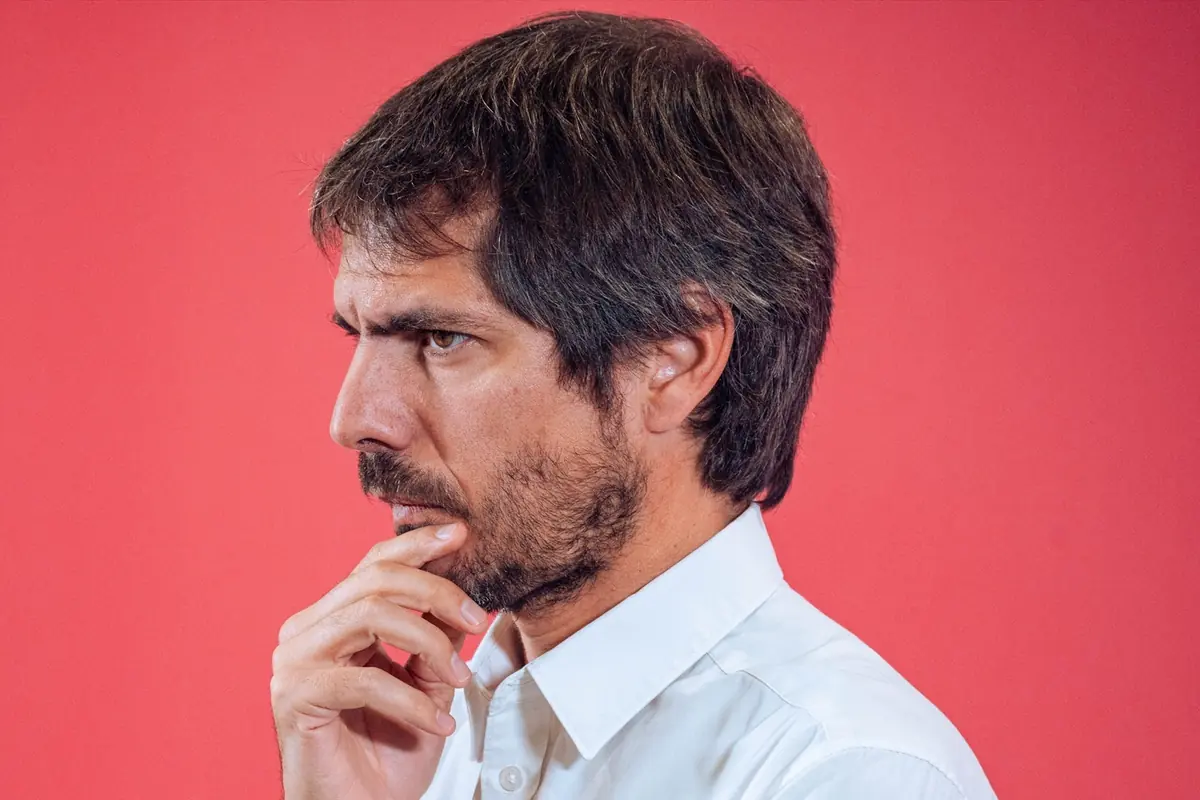 Hombre pensativo de perfil con camisa blanca tocándose la barbilla sobre fondo rojo