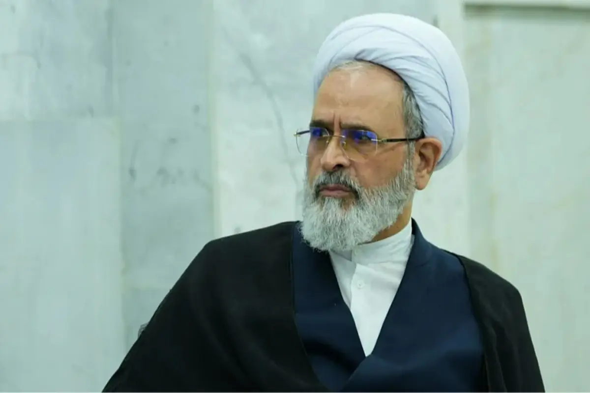 Hombre mayor con barba canosa gafas y turbante blanco vestido con túnica oscura frente a un fondo de mármol claro