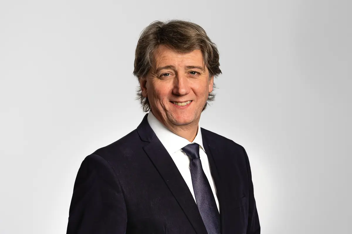 Retrato de un hombre de mediana edad con traje oscuro camisa blanca y corbata azul sonriendo frente a un fondo gris claro