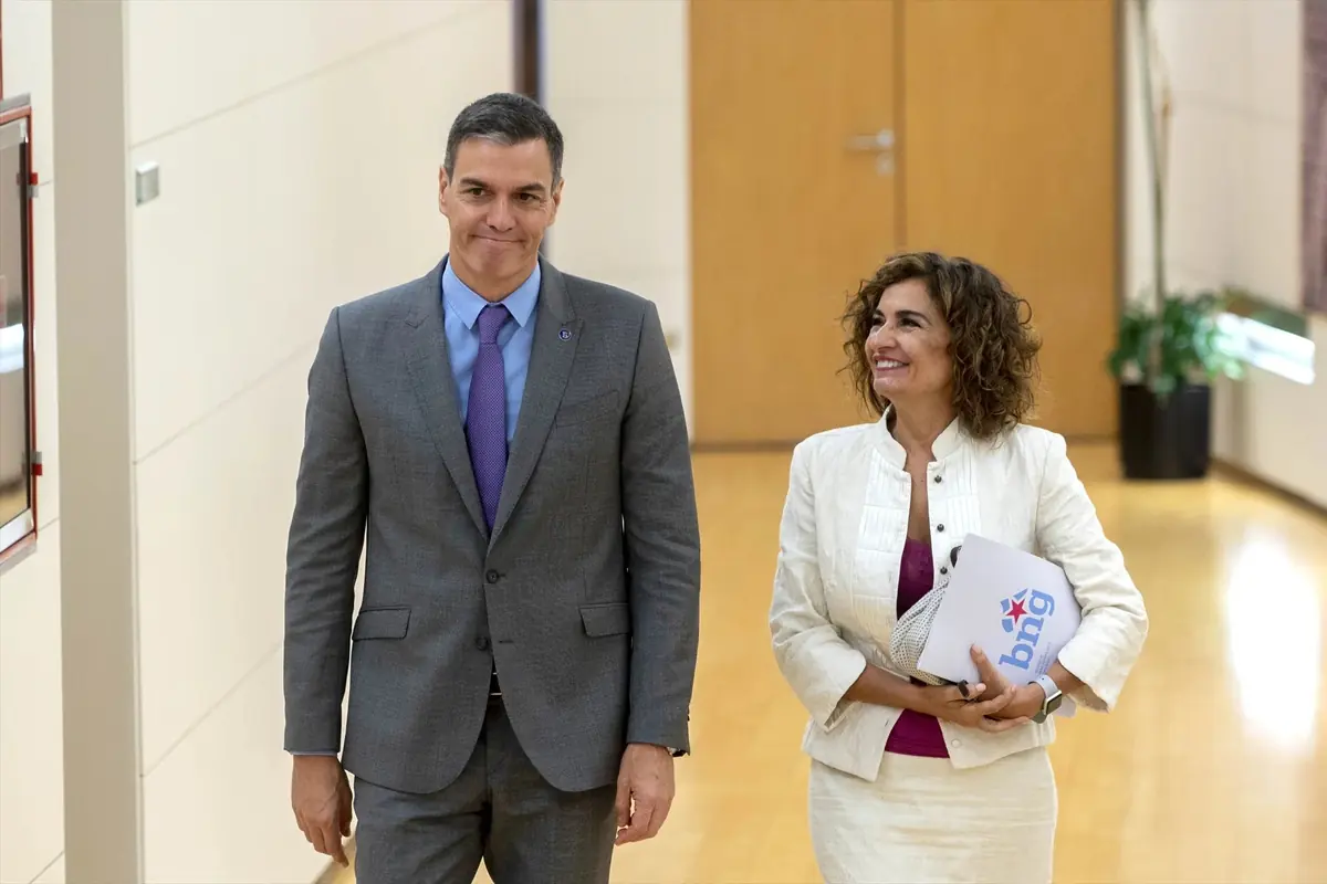 Hombre con traje gris y corbata morada camina junto a mujer con conjunto blanco que sostiene una carpeta en un pasillo de edificio institucional