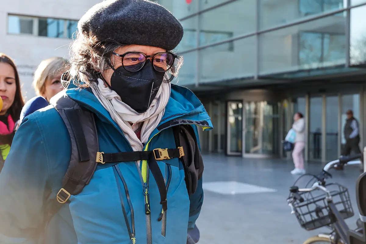 Persona mayor con boina gafas y mascarilla negra caminando con mochila y abrigo azul frente a un edificio moderno de cristal