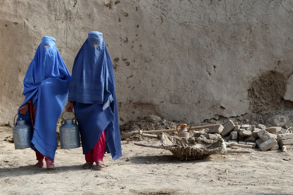 Dos mujeres con burka azul caminan por un camino de tierra llevando grandes bidones metálicos de agua junto a una pared de adobe agrietada Dos mujeres con burka azul caminan por un camino de tierra llevando grandes bidones metálicos de agua junto a una pared de adobe agrietada