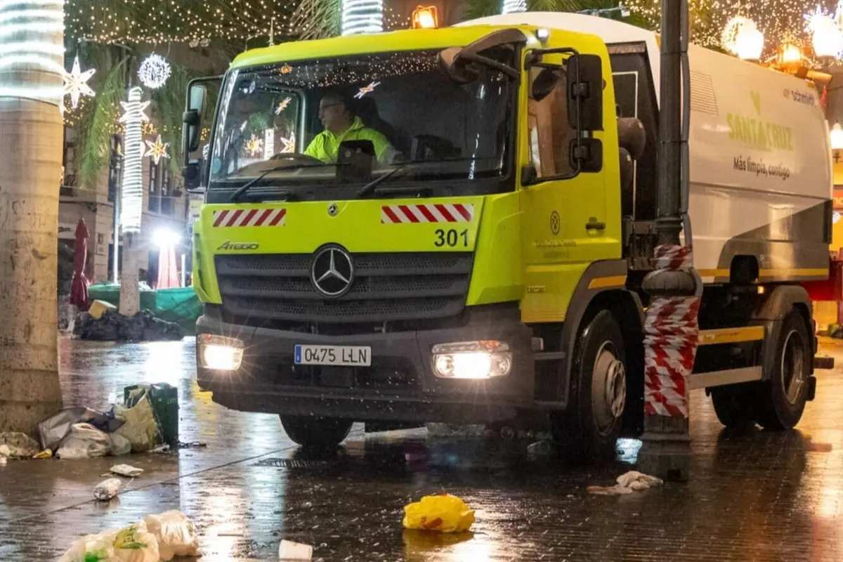 Camión de limpieza viaria trabajando de noche en una calle mojada y llena de basura con luces navideñas de fondo Camión de limpieza viaria trabajando de noche en una calle mojada y llena de basura con luces navideñas de fondo