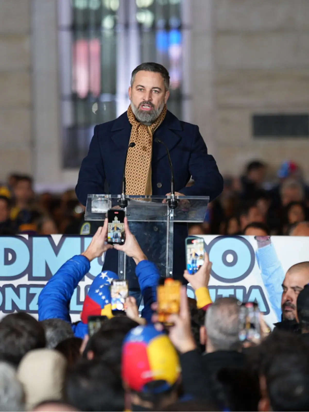 Hombre hablando en un atril transparente ante una multitud que lo fotografía con sus móviles en un acto público al aire libre