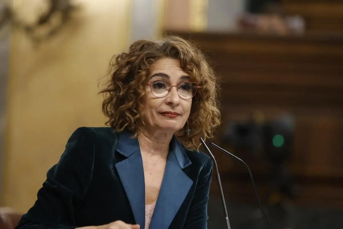 Mujer de mediana edad con gafas rojas y chaqueta verde oscuro hablando frente a un micrófono en un entorno institucional