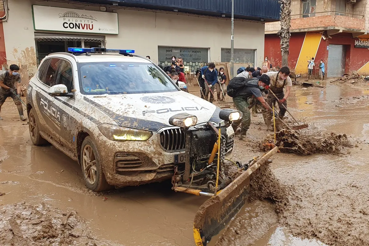 Coche de policía con pala frontal y voluntarios retirando barro en una calle inundada tras una riada