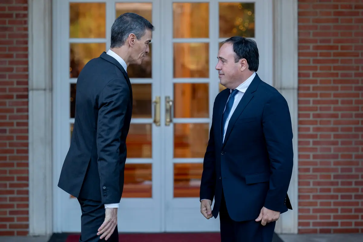Dos hombres con traje oscuro conversan frente a una puerta de cristal en un edificio de ladrillo