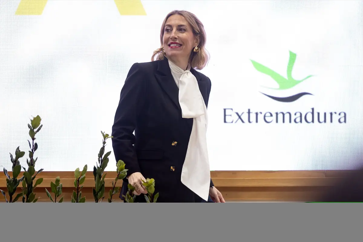 Mujer con traje negro y lazo blanco en el cuello sonriendo frente a un panel con el logotipo y la palabra Extremadura