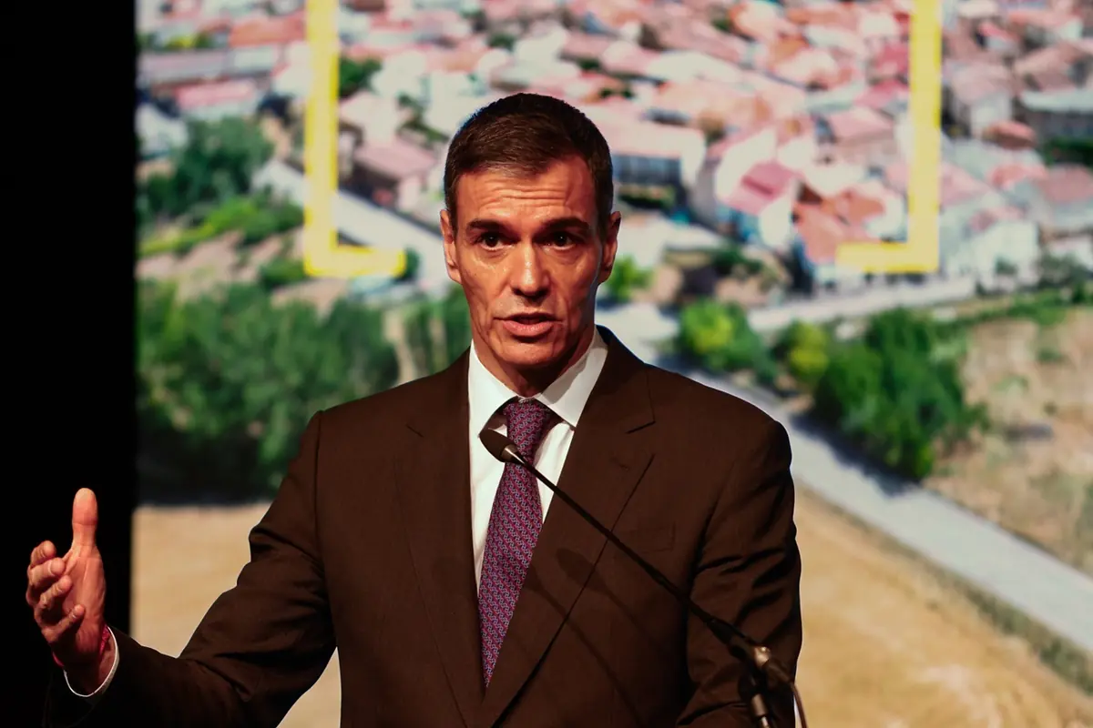 Hombre con traje oscuro y corbata hablando ante un micrófono con una imagen aérea de un pueblo desenfocada de fondo