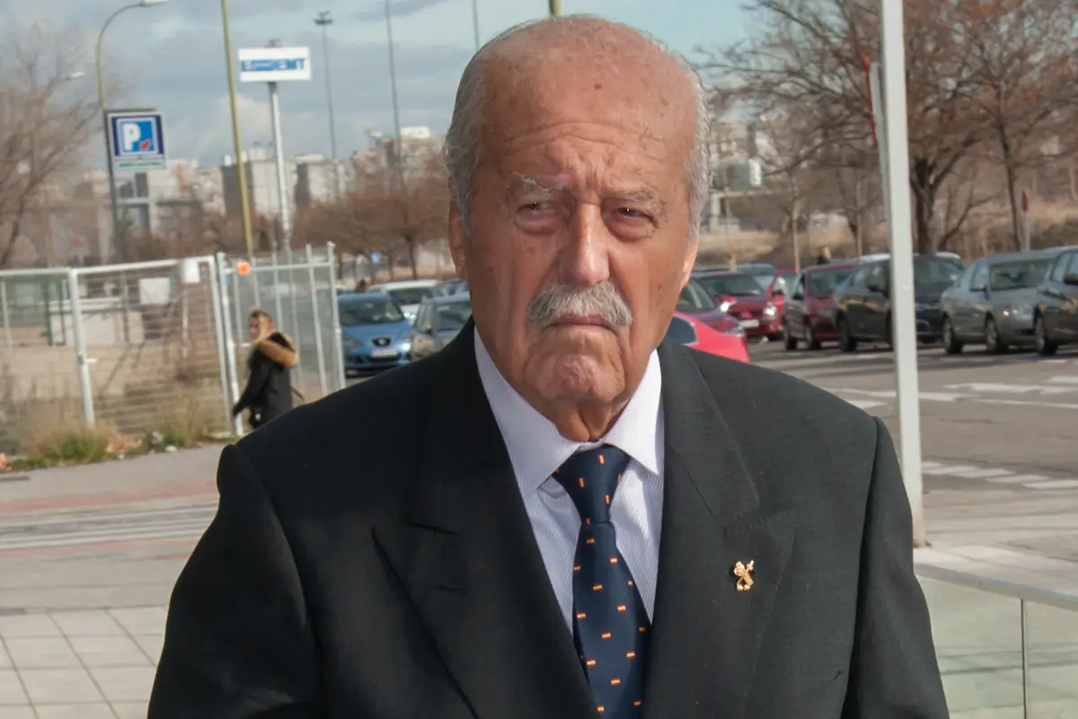 Hombre mayor con bigote y traje oscuro caminando por una acera en una zona urbana con coches aparcados al fondo