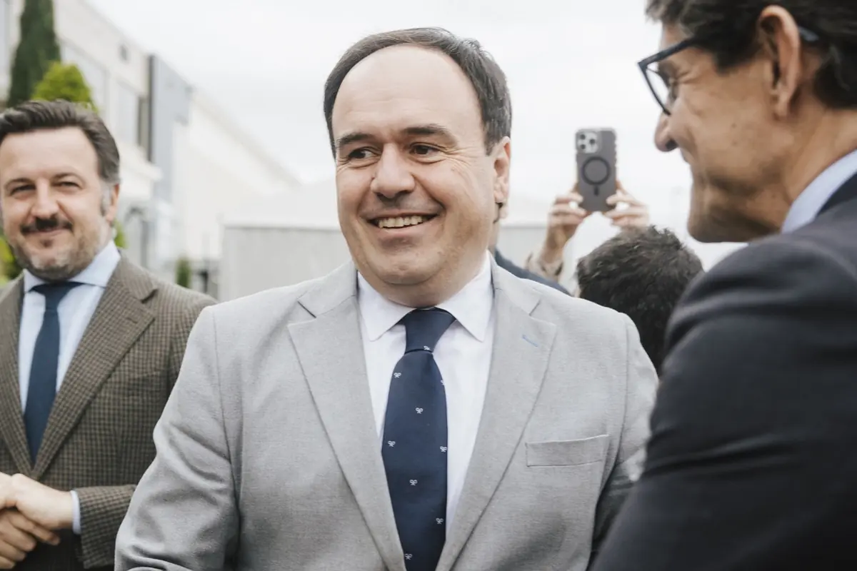 Hombre de mediana edad con traje gris y corbata azul sonriendo mientras conversa con otras personas en un evento al aire libre