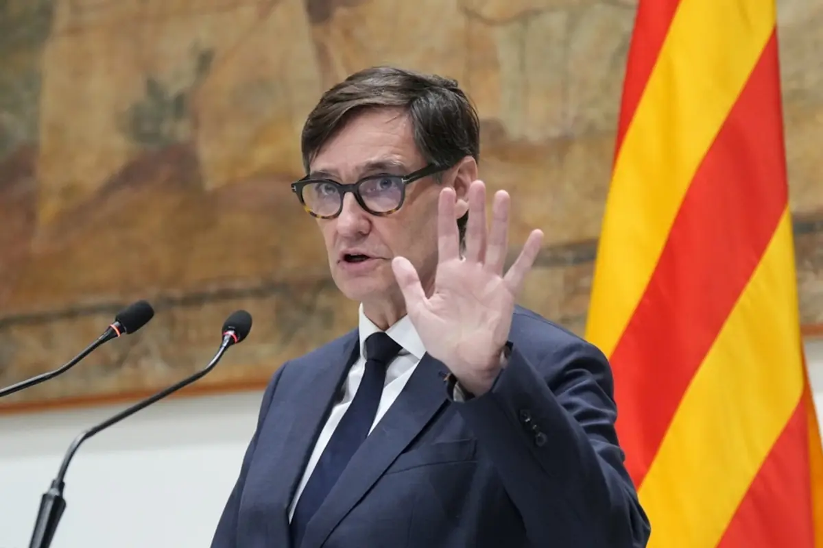Hombre con gafas y traje oscuro hablando en un atril con micrófonos y la bandera catalana al fondo mientras levanta la mano