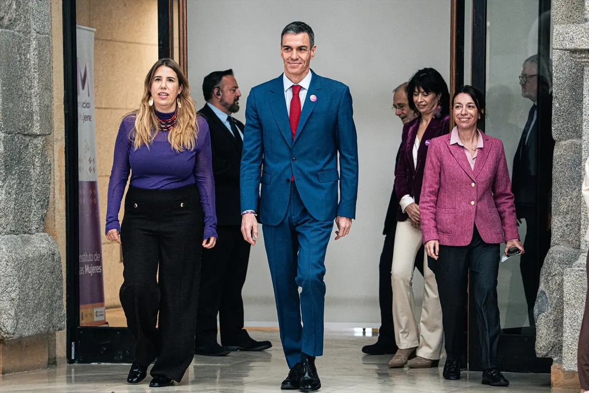 Grupo de personas con ropa formal saliendo de un edificio encabezadas por un hombre con traje azul y corbata roja acompañado por dos mujeres con chaquetas de colores vivos