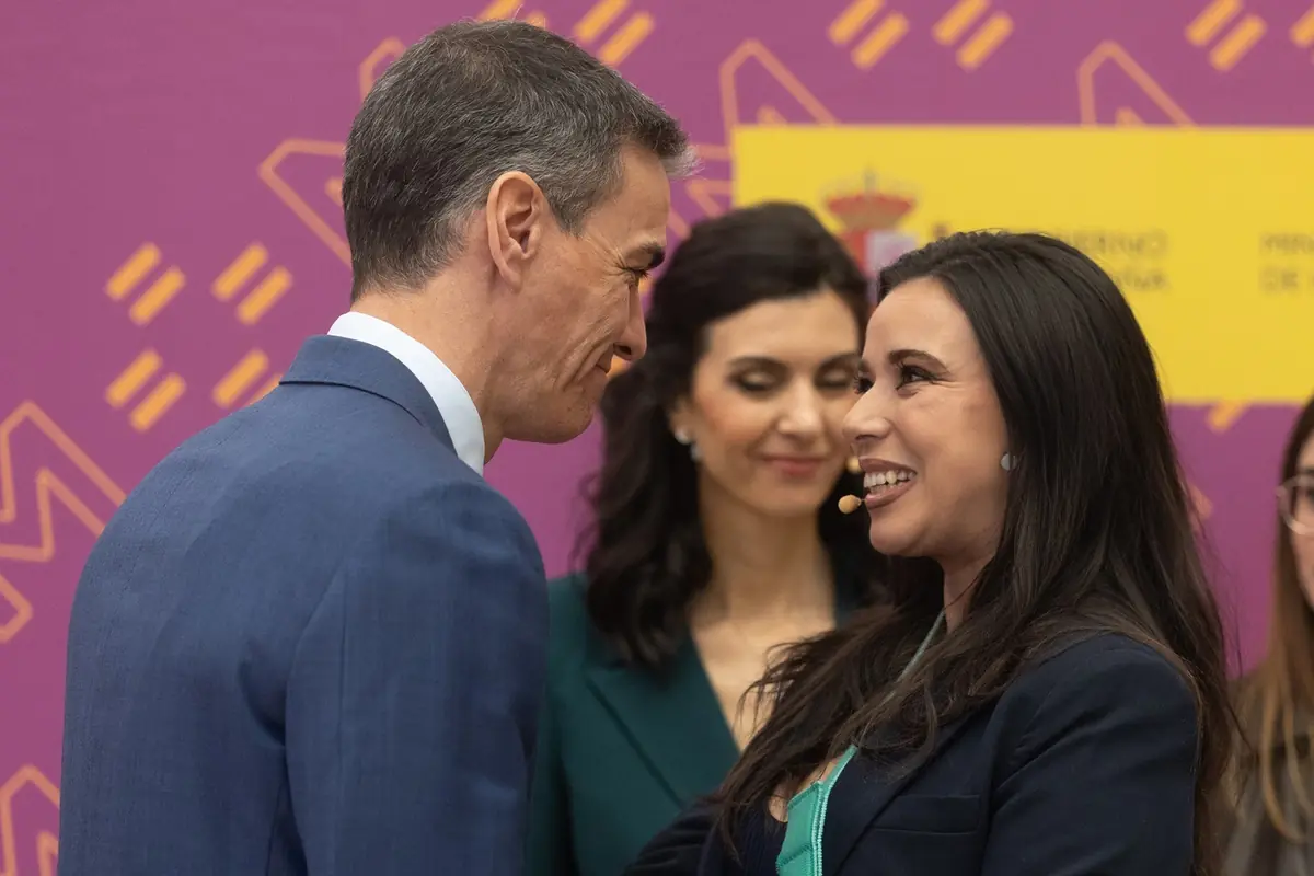 Hombre y mujer con micrófonos de diadema se saludan sonrientes en un acto público con fondo morado y amarillo