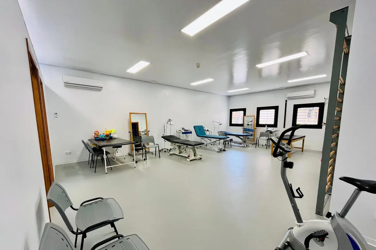 Sala amplia y luminosa de fisioterapia con camillas, mesas de trabajo, sillas, bicicleta estática y equipamiento médico distribuido en un entorno blanco y ordenado