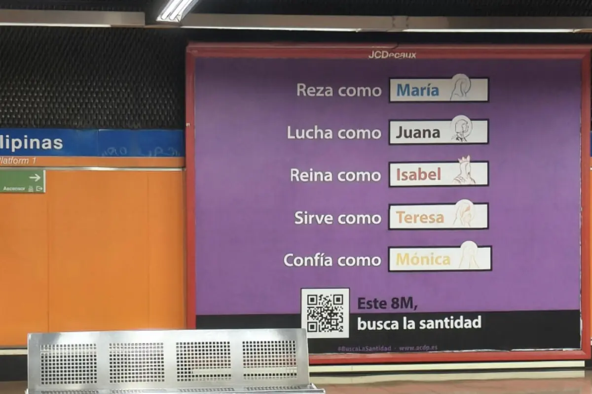 Cartel publicitario de fondo morado en una estación de metro con el texto Reza como María Lucha como Juana Reina como Isabel Sirve como Teresa Confía como Mónica y el eslogan Este 8M busca la santidad junto a un código QR