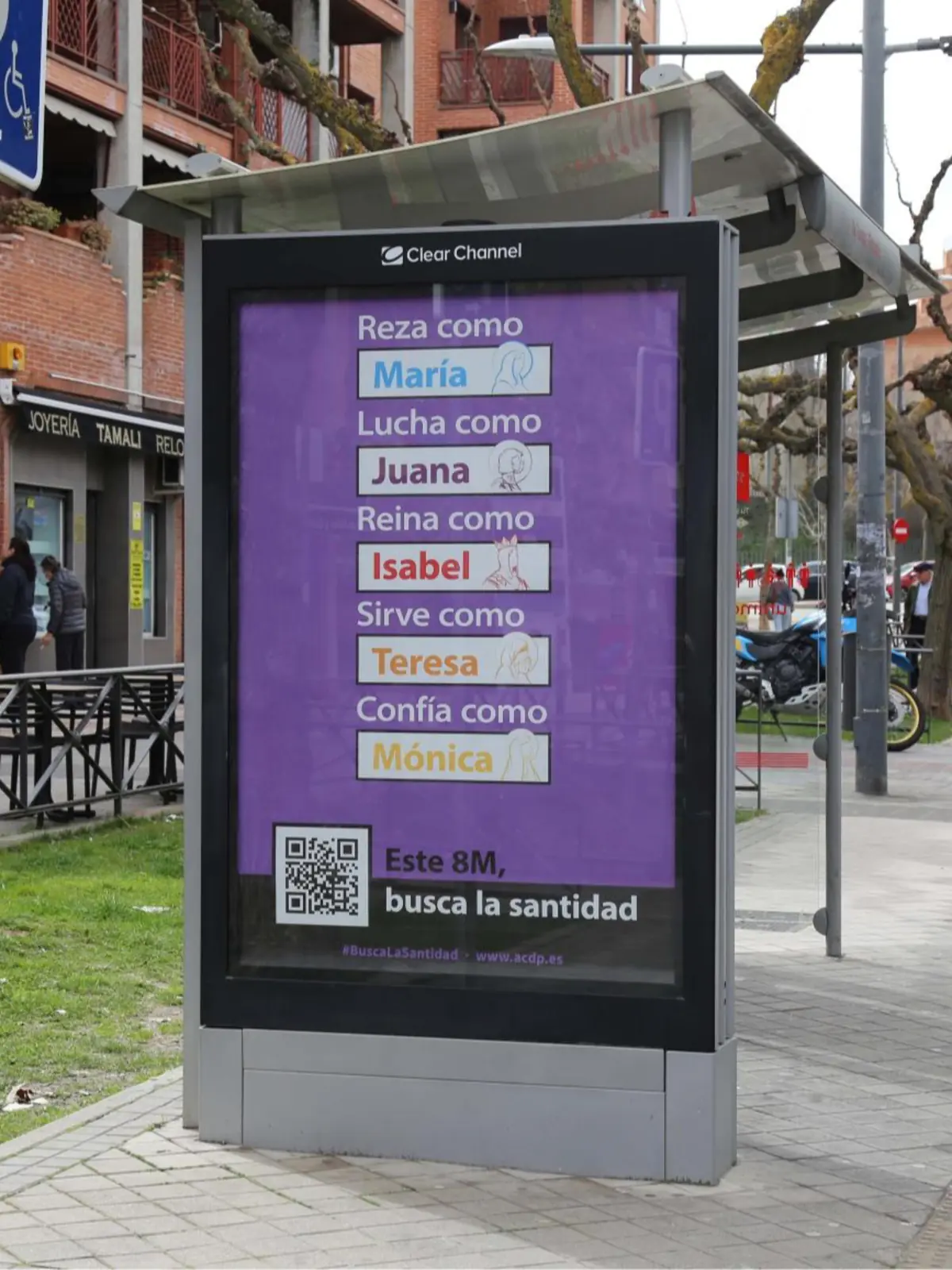 Marquesina de autobús en una calle con un cartel publicitario morado que muestra mensajes religiosos sobre mujeres con textos como Reza como María Lucha como Juana Reina como Isabel Sirve como Teresa Confía como Mónica y el lema Este 8M busca la santidad junto a un código QR