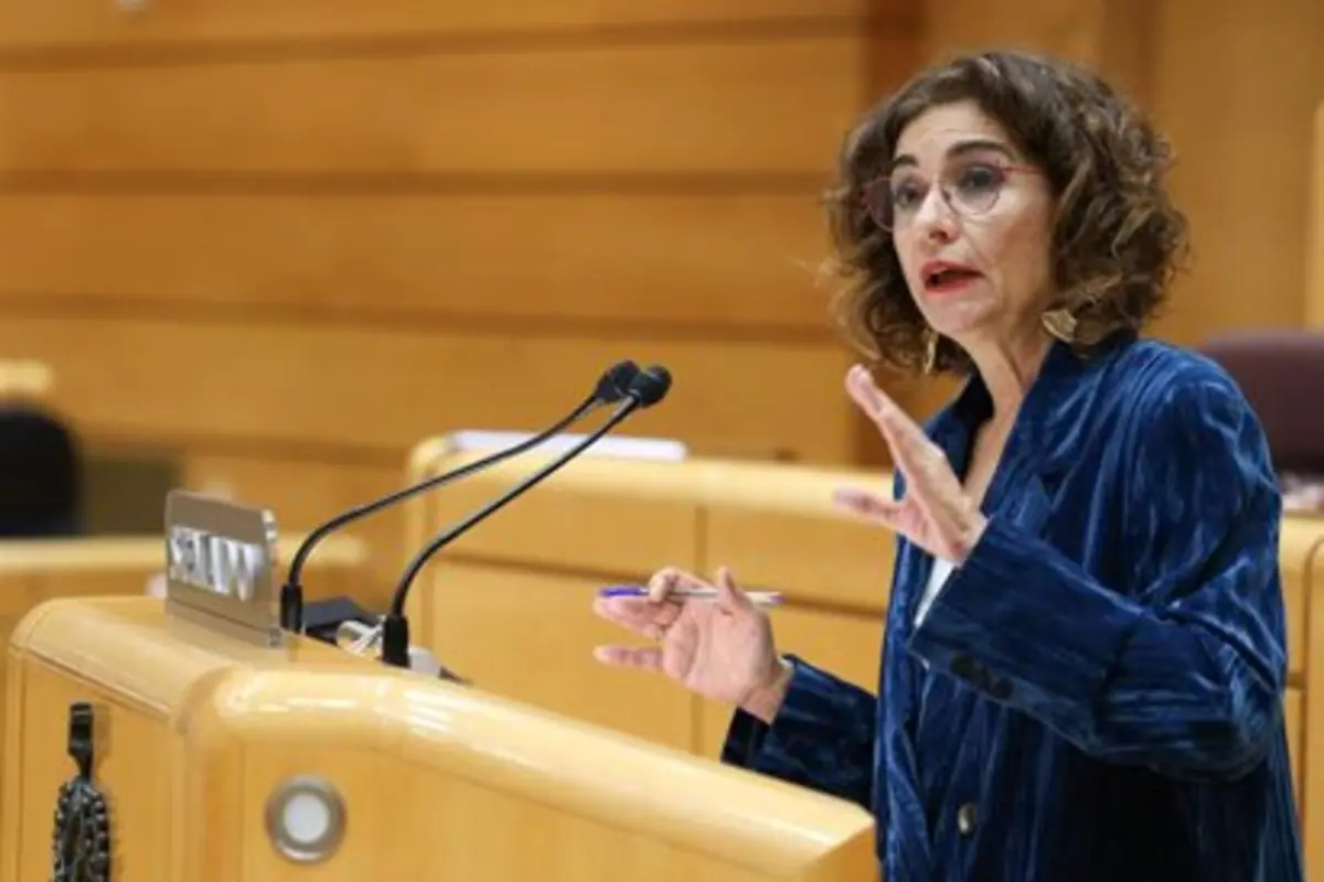 Mujer con gafas y chaqueta azul hablando gesticulando detrás de un atril con micrófonos en un salón de plenos