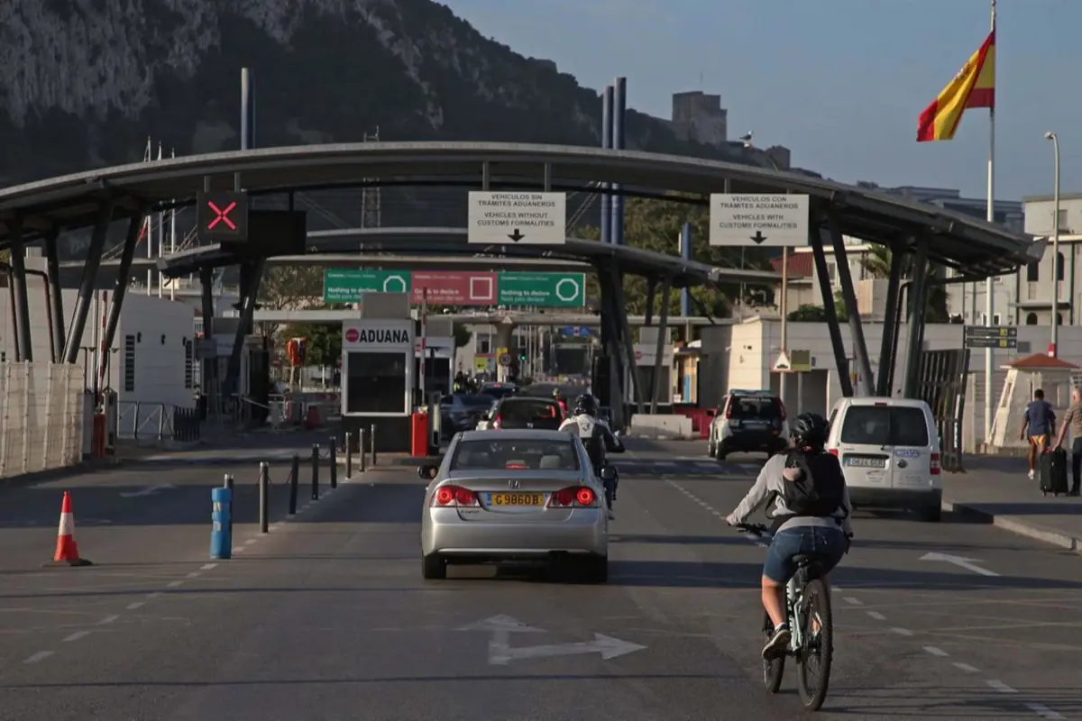 Frontera por carretera con cabinas de aduana y varios vehículos y ciclistas entrando en España bajo una gran estructura metálica y una bandera española ondeando a la derecha