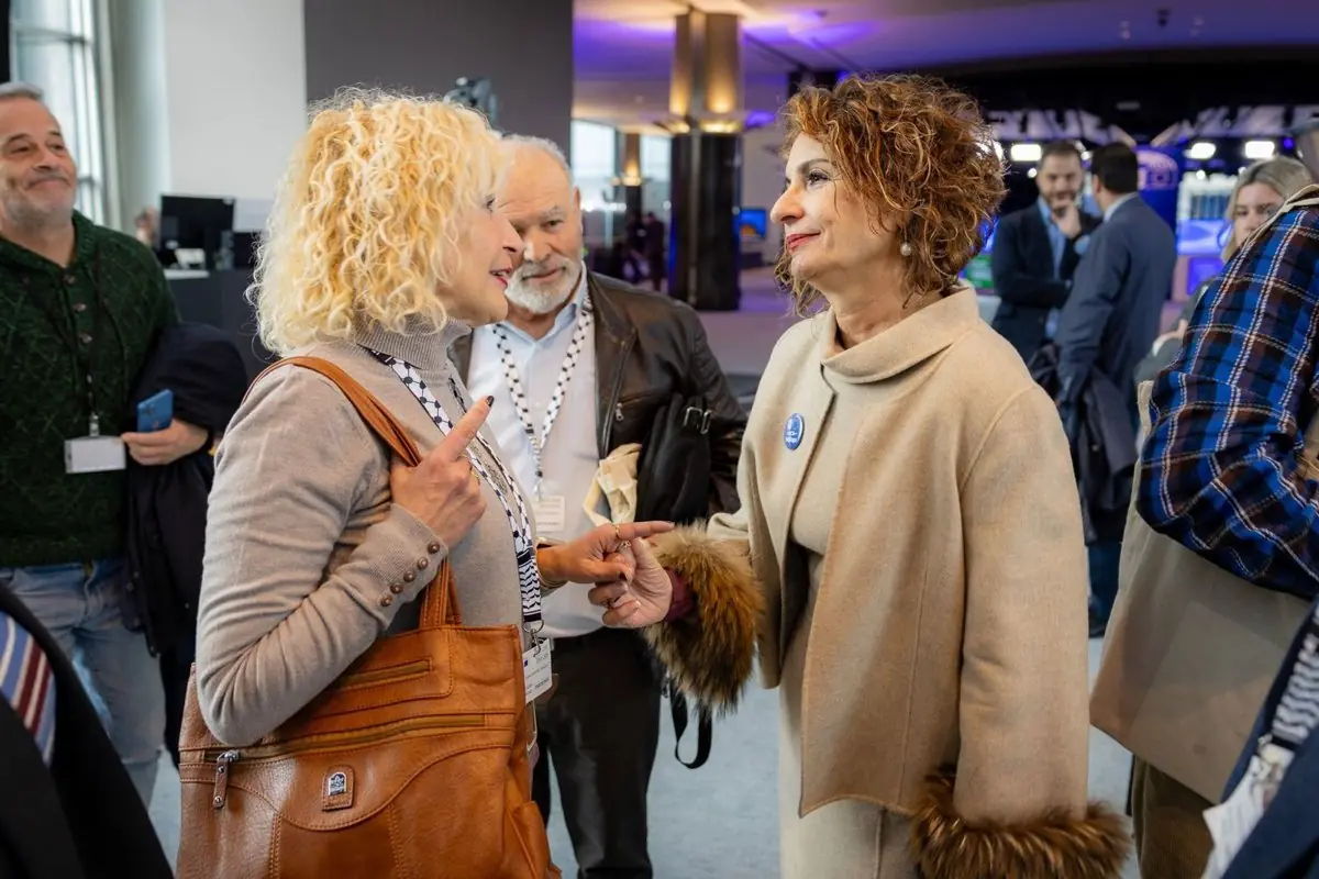 Dos mujeres conversan de cerca en un evento profesional rodeadas de otras personas en un espacio interior moderno