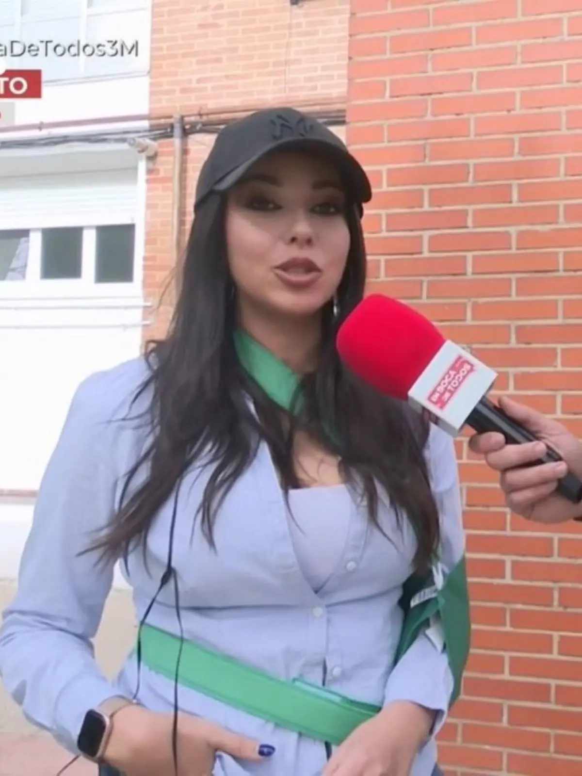Mujer joven con gorra negra y camisa azul siendo entrevistada en la calle frente a una pared de ladrillo mientras sostiene un micrófono rojo cerca de su rostro