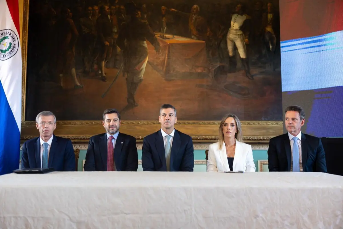 Cinco personas con traje formal sentadas detrás de una mesa en un salón oficial con un gran cuadro histórico y una bandera de Paraguay al fondo
