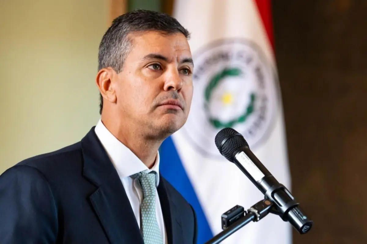 Hombre de traje hablando frente a un micrófono con una bandera de Paraguay de fondo