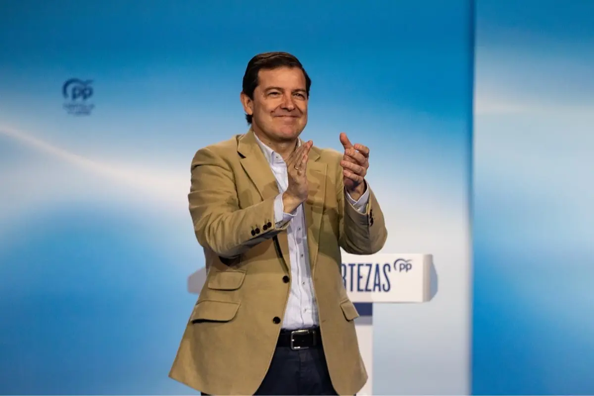 Hombre con americana beige aplaudiendo sobre un escenario azul junto a un atril con el logotipo del PP Hombre con americana beige aplaudiendo sobre un escenario azul junto a un atril con el logotipo del PP