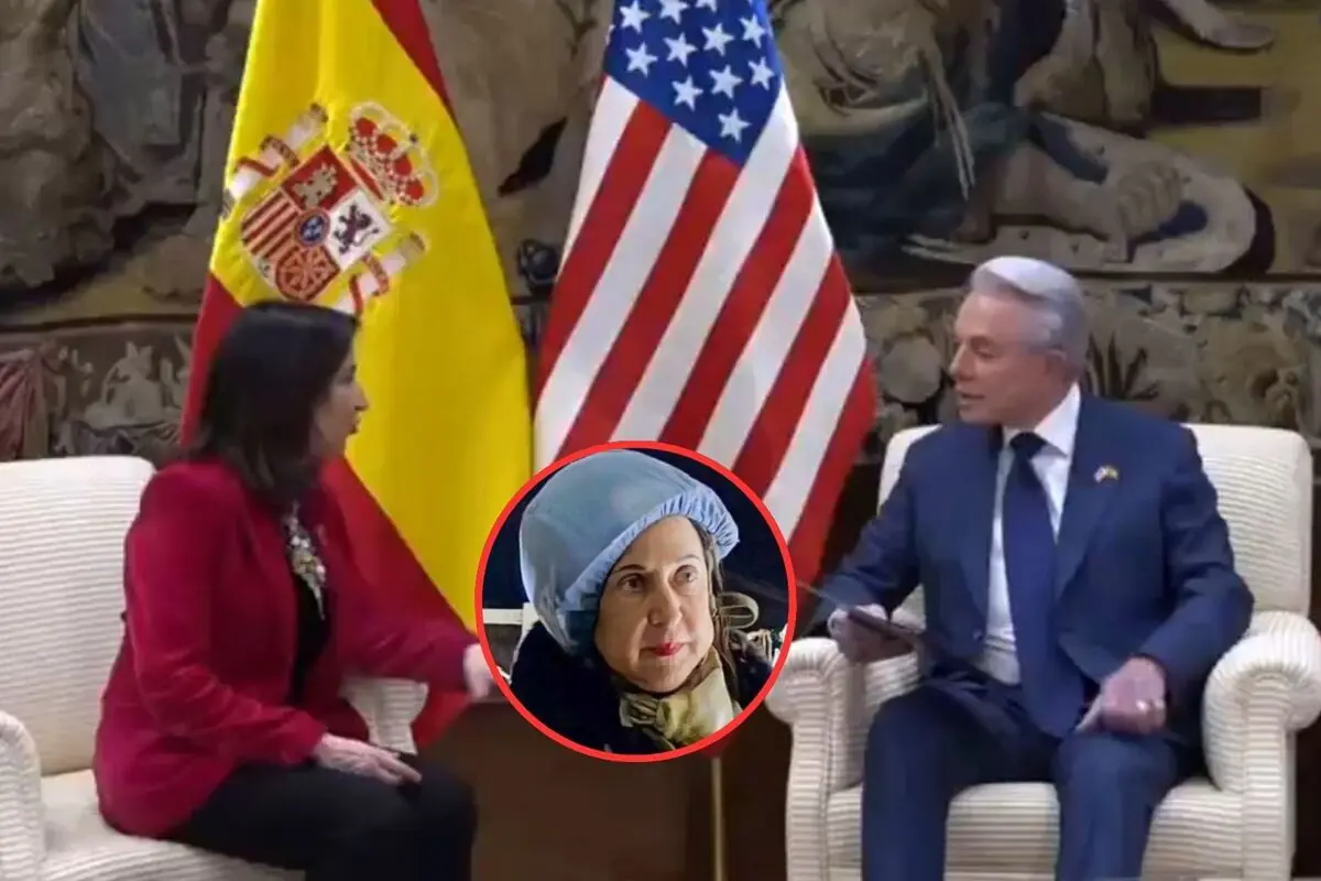 Reunión entre una mujer con chaqueta roja y un hombre con traje azul sentados frente a las banderas de España y Estados Unidos con un recuadro circular que muestra el rostro de una mujer mayor con gorro azul