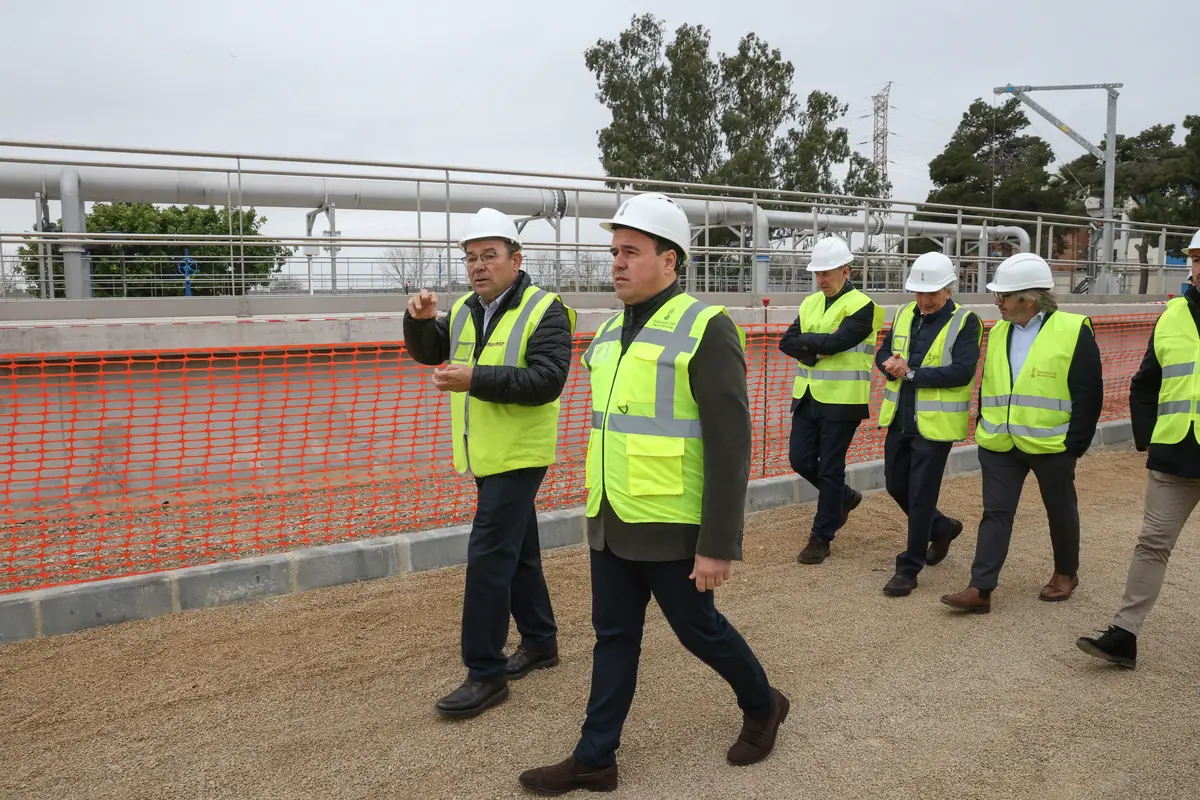 Grupo de hombres con chalecos reflectantes y cascos blancos recorriendo una obra de infraestructura junto a una valla de seguridad naranja Grupo de hombres con chalecos reflectantes y cascos blancos recorriendo una obra de infraestructura junto a una valla de seguridad naranja