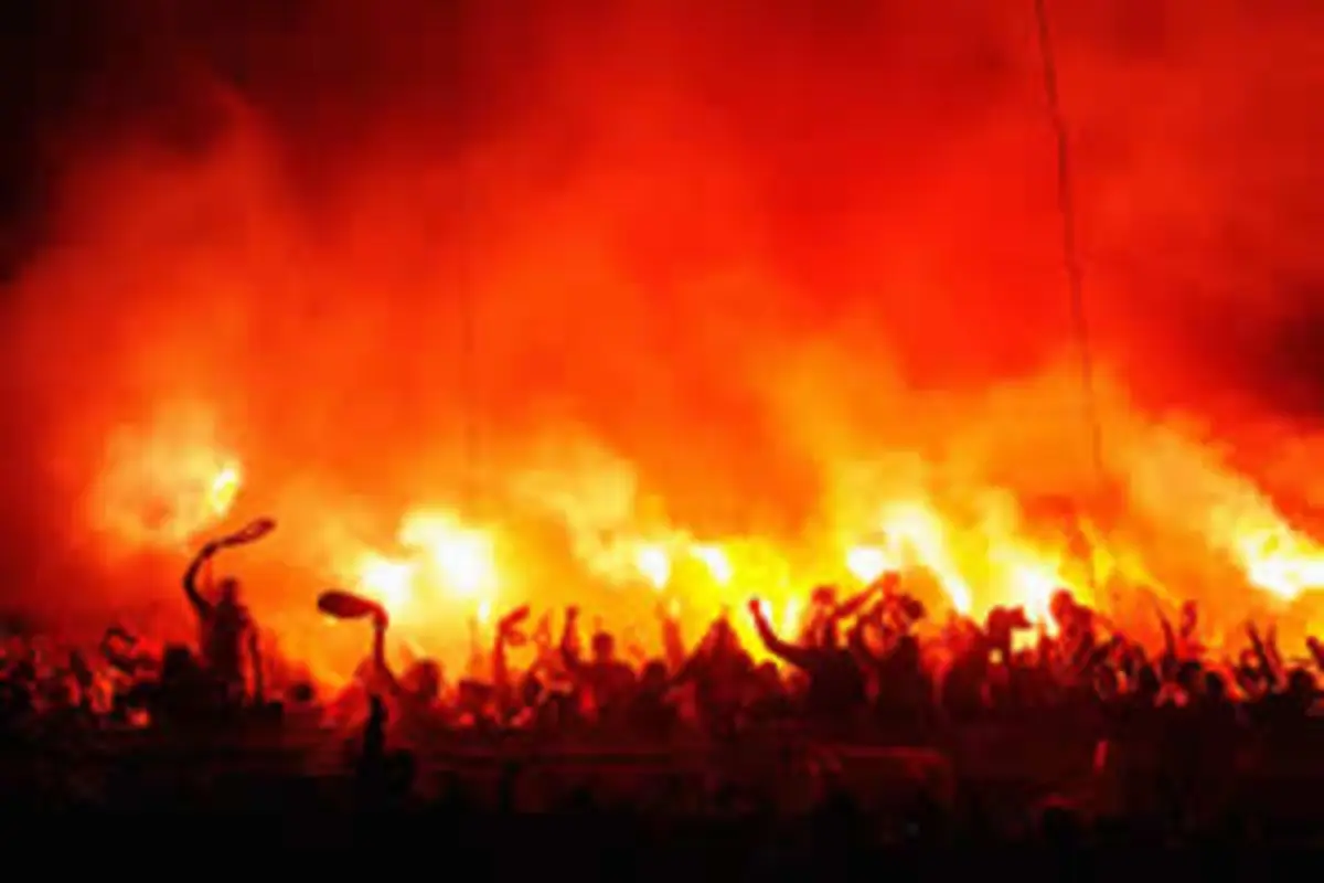 Multitud de aficionados celebrando de noche con bengalas rojas encendidas y humo denso en el estadio