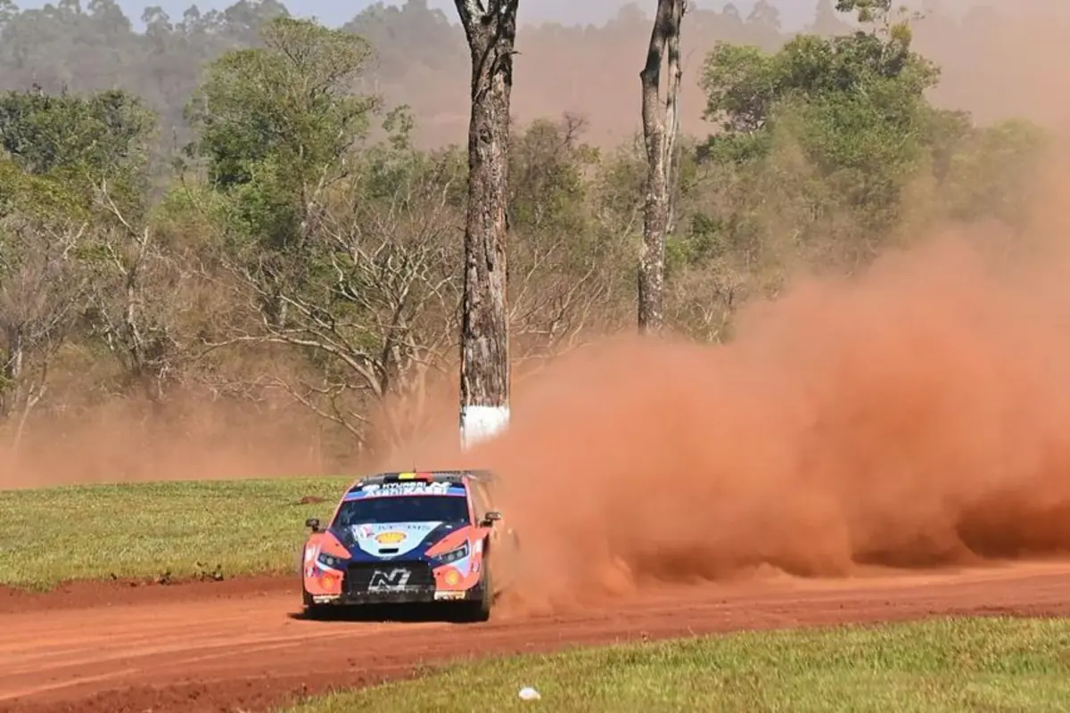 Auto de rally avanzando a gran velocidad por un camino de tierra roja levantando una gran nube de polvo en un entorno de bosque verde