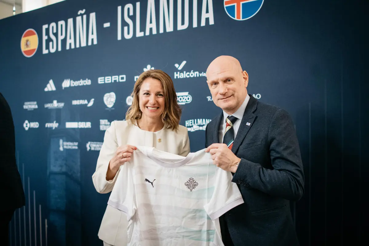 Dos representantes de España e Islandia posan sonrientes sosteniendo una camiseta de fútbol blanca frente a un panel con el texto España Islandia y logotipos de patrocinadores