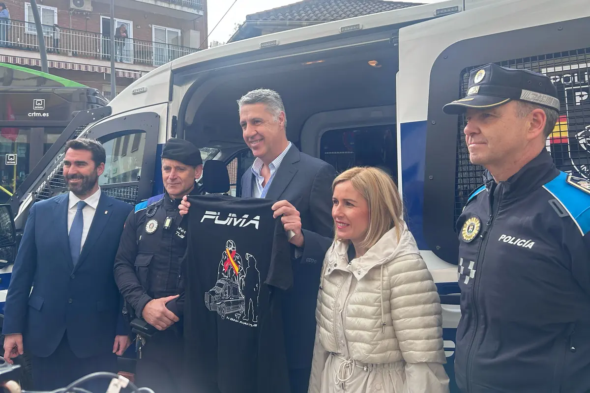 Grupo de autoridades y agentes de policía posando frente a una furgoneta policial mientras un hombre sostiene una camiseta negra con el logo PUMA y un diseño de policías y vehículo patrulla