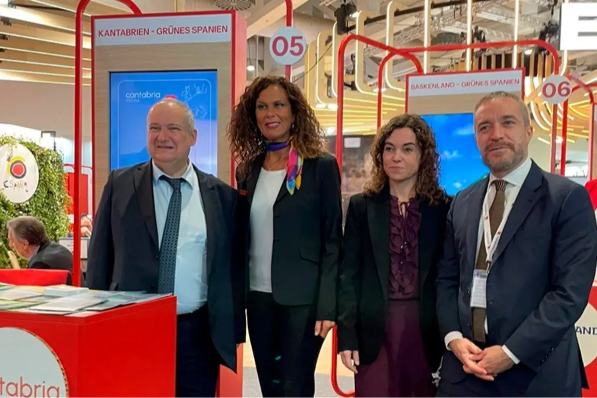 Cuatro personas con traje posan sonrientes en un stand de feria turística de España con carteles de Cantabria y otras regiones de “Grünes Spanien” al fondo