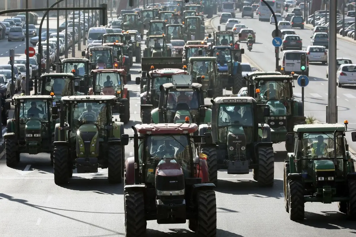 Fila de tractores ocupando varios carriles de una avenida urbana durante una protesta agrícola rodeados de coches en ambos lados