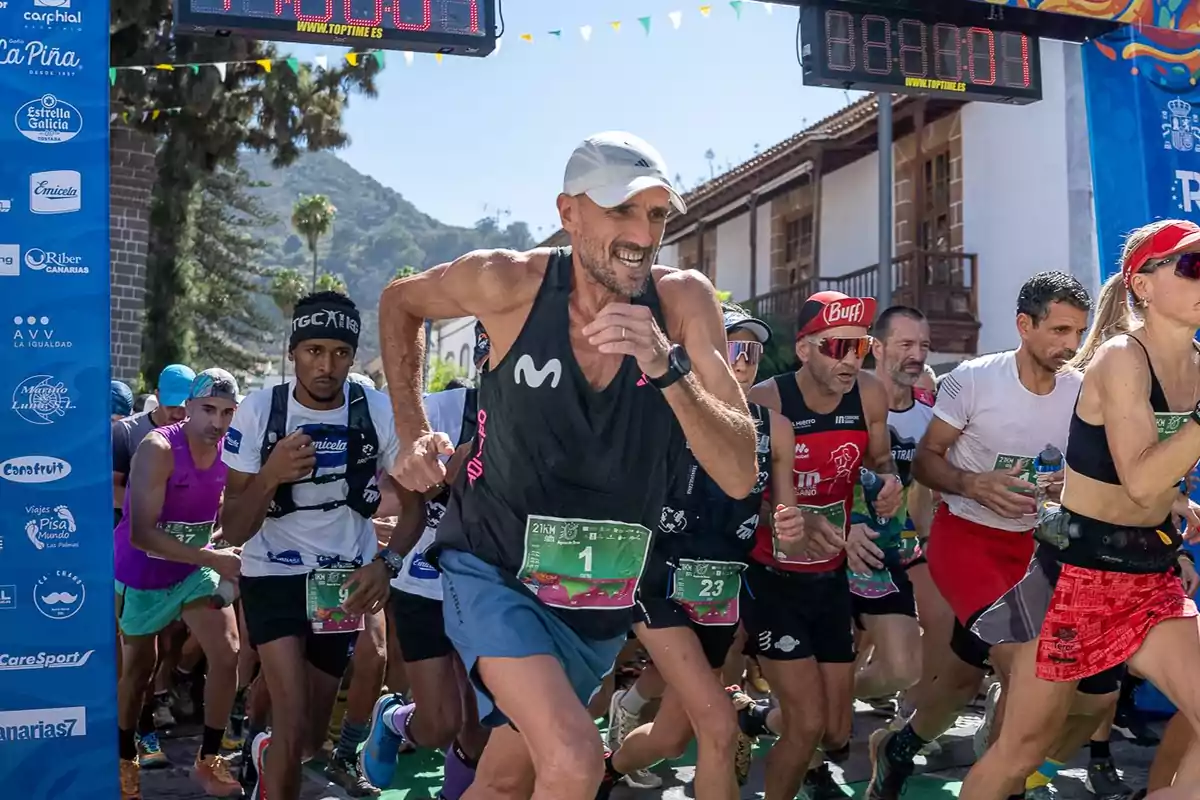 Corredores participando en una carrera al aire libre, con un hombre en primer plano llevando una gorra blanca y una camiseta sin mangas negra, rodeado de otros corredores con diferentes atuendos deportivos y números de identificación.