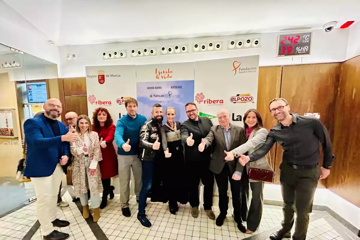 A group of people posing together in front of a background with logos and posters, all giving thumbs-up gestures.