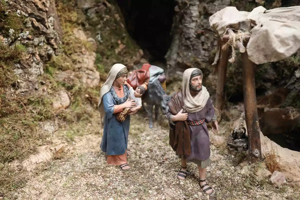 Una escena de un belén con figuras que representan a una mujer sosteniendo a un bebé y un hombre caminando junto a un burro en un entorno natural.