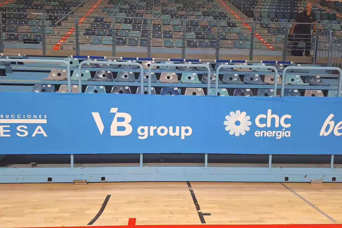 Gradas vacías de un estadio con una valla publicitaria azul que muestra varios logotipos de empresas.