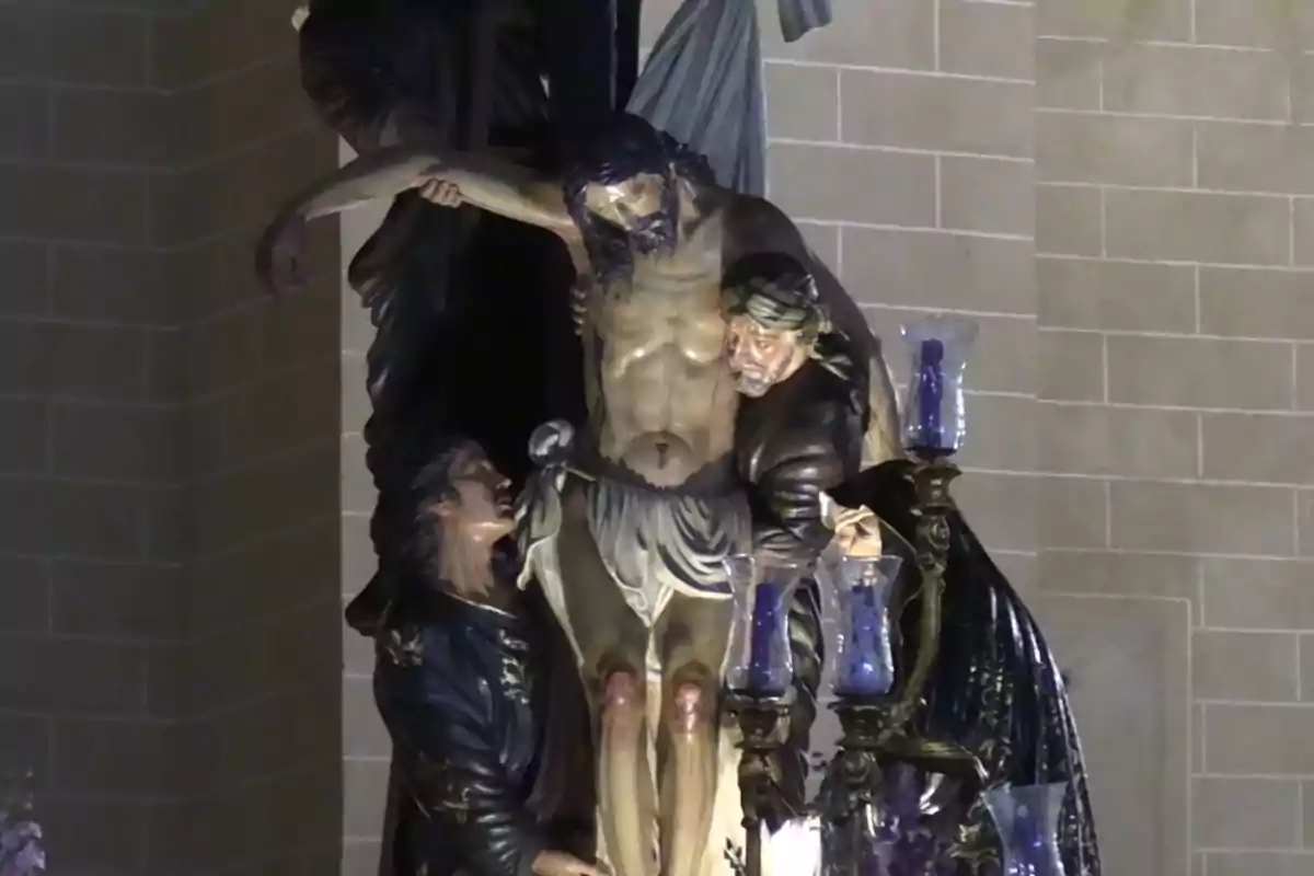 Escultura religiosa que representa el descendimiento de Cristo de la cruz con figuras humanas sosteniéndolo en un entorno de iluminación tenue. Escultura religiosa que representa el descendimiento de Cristo de la cruz con figuras humanas sosteniéndolo en un entorno de iluminación tenue.