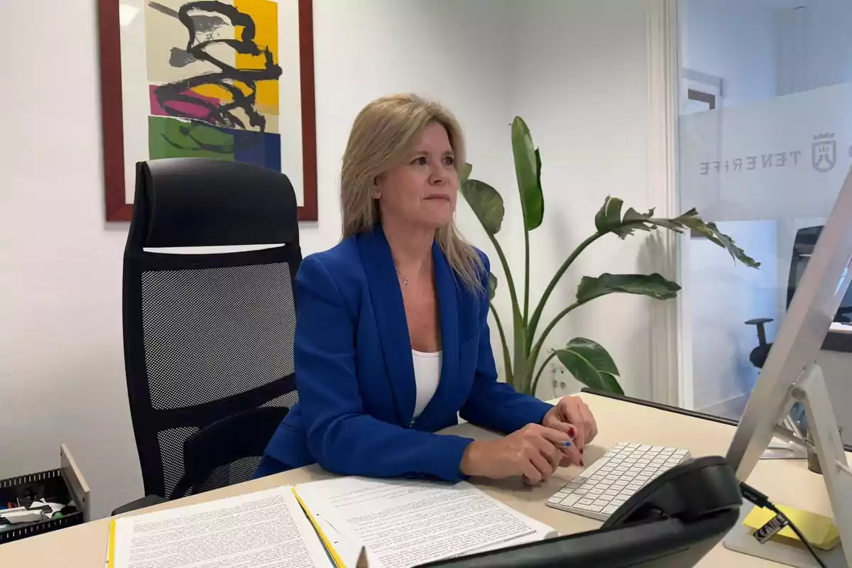 Mujer trabajando en una oficina frente a una computadora con documentos sobre el escritorio y una planta al fondo.